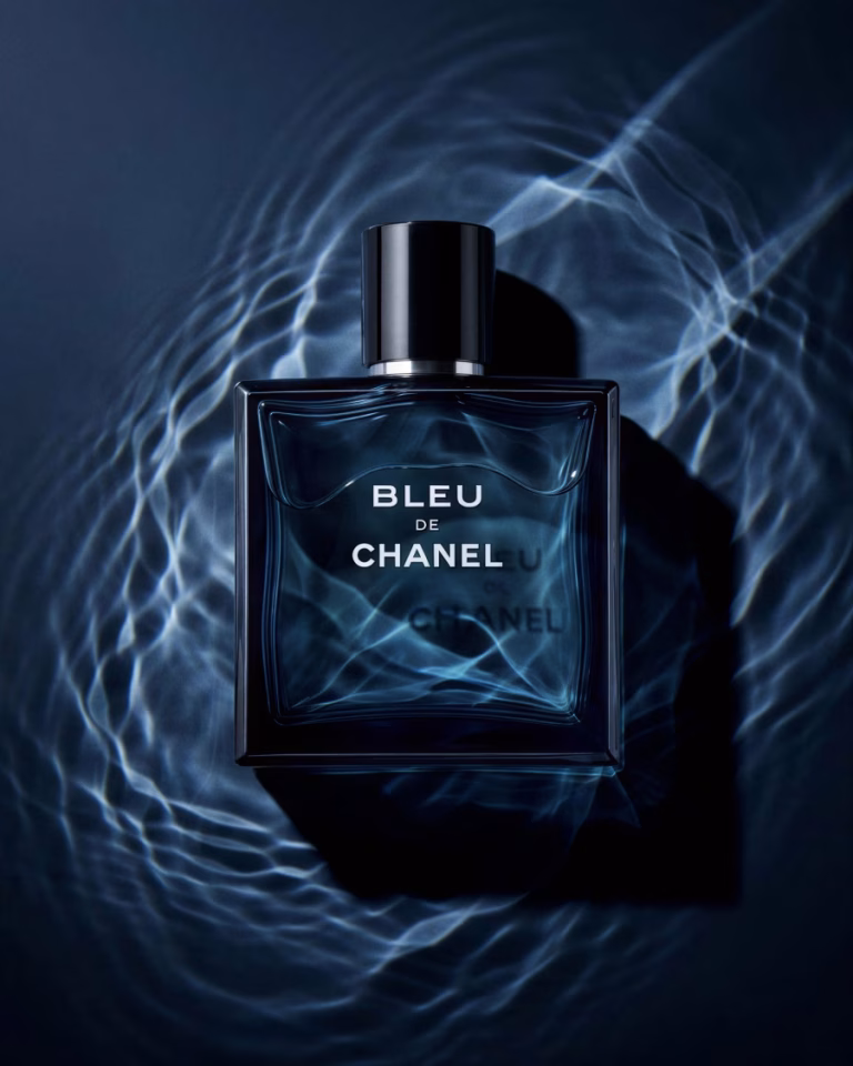 Flacon de parfum carré en verre bleu profond, capot noir, posé à plat dans des ondes d'eau lumineuses sur fond sombre — mise en scène studio