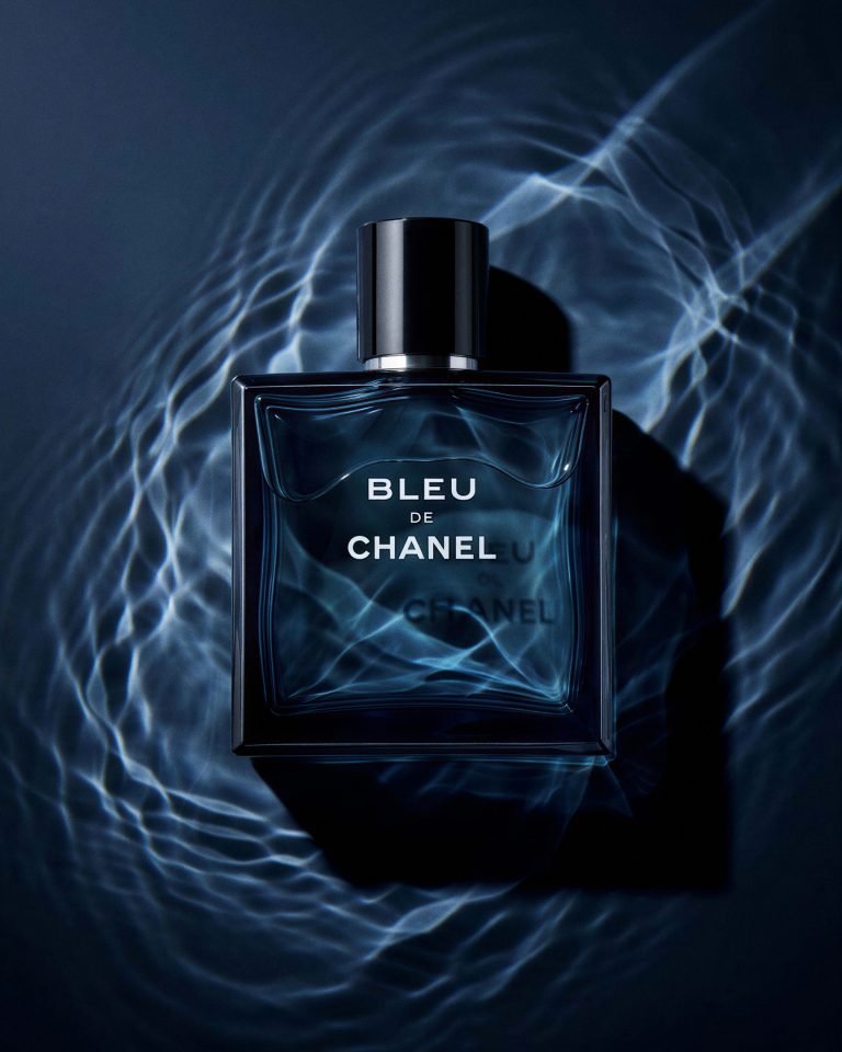 Flacon de parfum carré en verre bleu profond, capot noir, posé à plat dans des ondes d'eau lumineuses sur fond sombre — mise en scène studio
