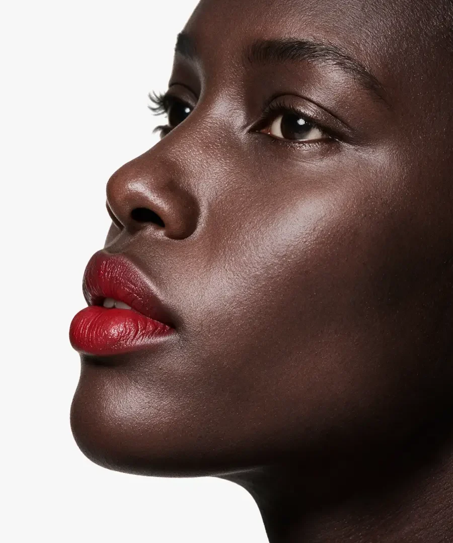 Portrait beauté en gros plan, vue de profil : une femme au teint mat, rouge à lèvres rouge intense au fini mat légèrement dégradé sur le contour, peau lumineuse, fond blanc épuré.